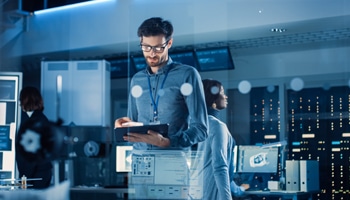 In Technology Research Facility: Chief Engineer Stands in the Middle of the Lab and Uses Tablet Computer. Team of Industrial Engineers, Developers Work on Engine Design Use Digital Whiteboard and Computers